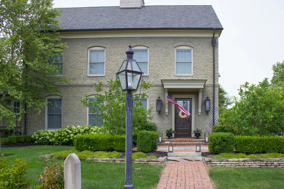 Front approach to estate home with brick walk, lamp post, trimmed hedges, lawn, and stone architecture.